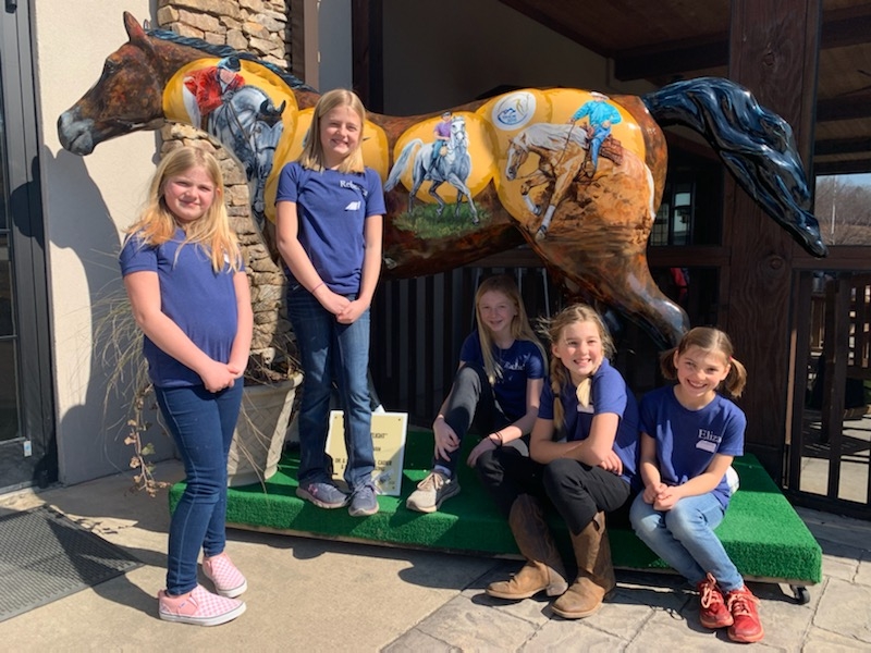 A group of children in front of a statue painted with scenes of horsemanship.